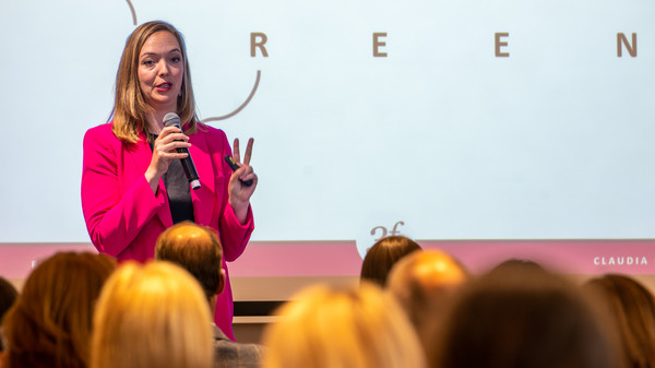 Frau im pinken Blazer spricht mit Mikrophon vor Teilnehmenden