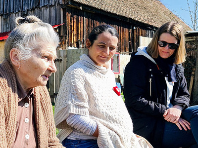 Drei Frauen unterschiedlichen Alters sitzen vor einem Holzhaus