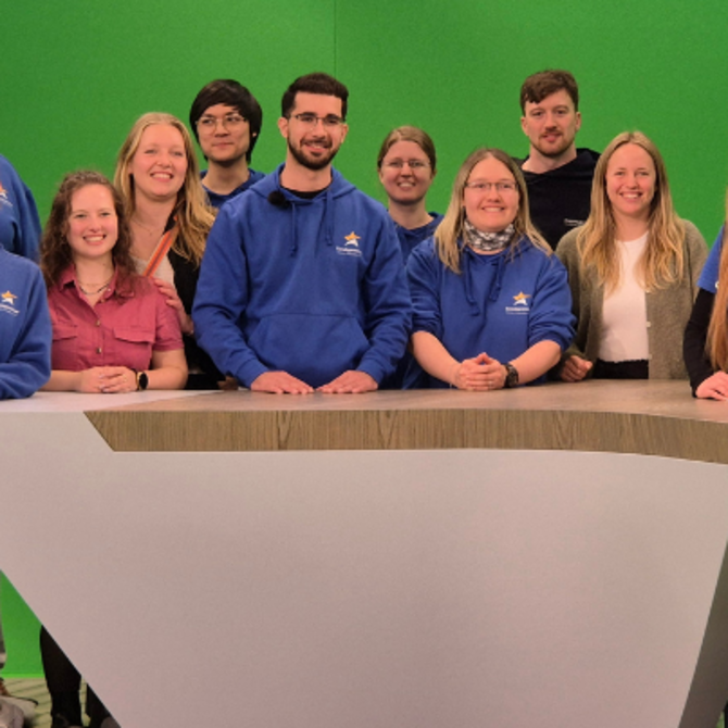 Teilnehmende des Treffens stehen vor einem Rednerpult in einem TV-Studio mit Greenscreen-Hintergrund.