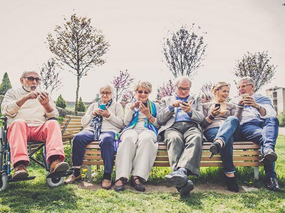 Gruppe älterer Menschen mit Smartphones auf einer Bank im Park: Erasmus+ Partnerschaft entwickelt Trainingsprogramm für Ältere zur Nutzung von Assistenztechnologie