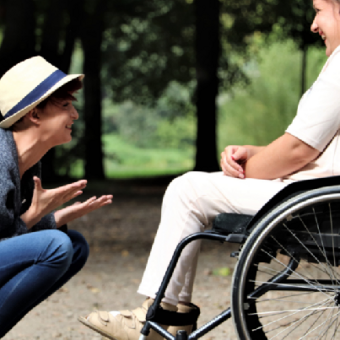 junger Mann kniet vor junger Frau im Rollstuhl und lacht mit ihr