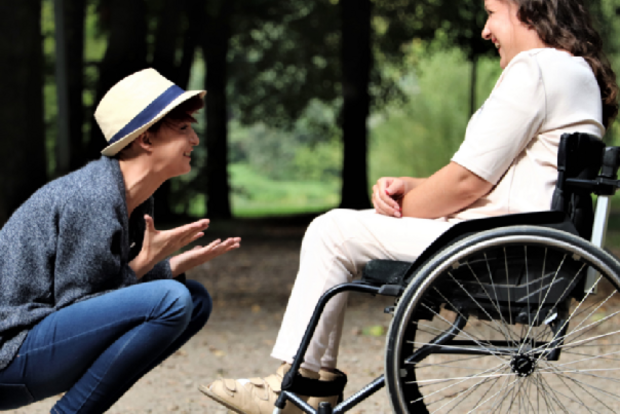 Ein junger Mann hockt vor einer jungen Frau im Rollstuhl und lacht mit ihr