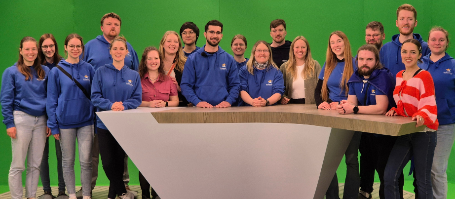 Teilnehmende des Treffens stehen vor einem Rednerpult in einem TV-Studio mit Greenscreen-Hintergrund.