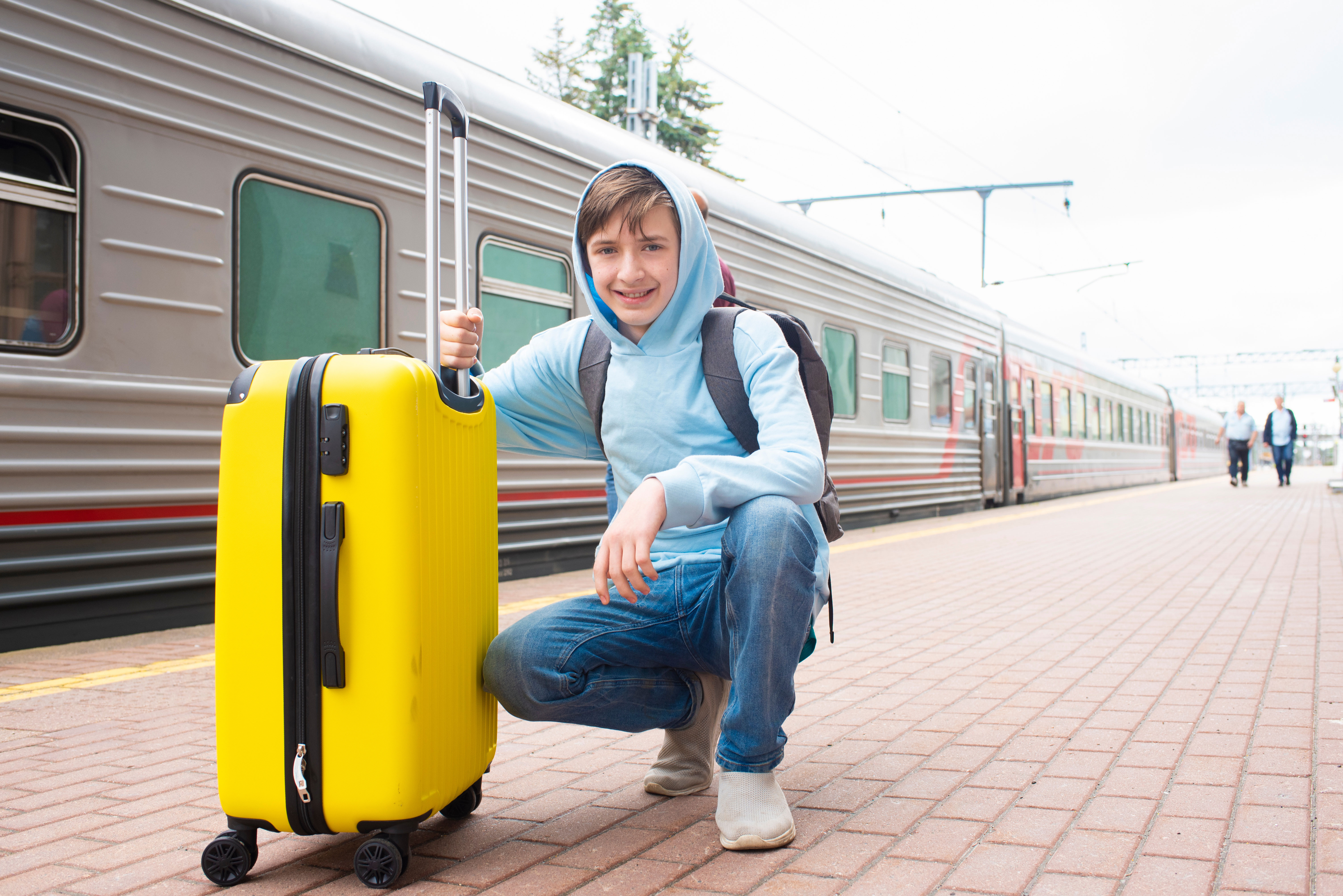 Ein etwa 12-j&auml;hriger Junge mit einem gelben Koffer hockt auf dem Bahnsteig in der N&auml;he des Zuges.