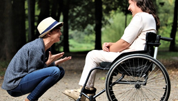 Zwei Frauen lachen sich an. Eine sitzt in der Hocke, die andere im Rollstuhl. Sie befinden sich auf einem Waldweg.