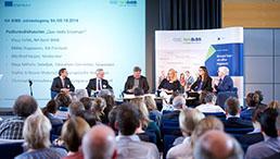 Sechs Menschen sitzen auf einem Podium, im Vordergrund sieht man die Hinterköpfe der Zuschauer