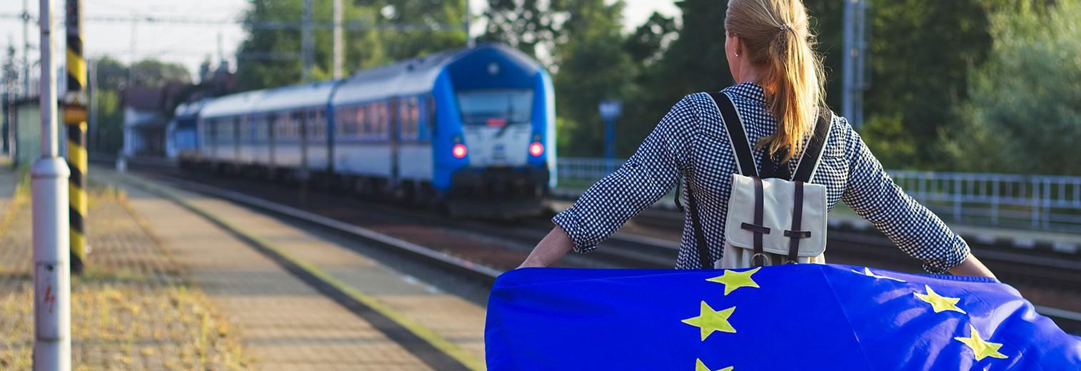 Eine Junge Frau steht mit der Europaflagge am Bahnhof